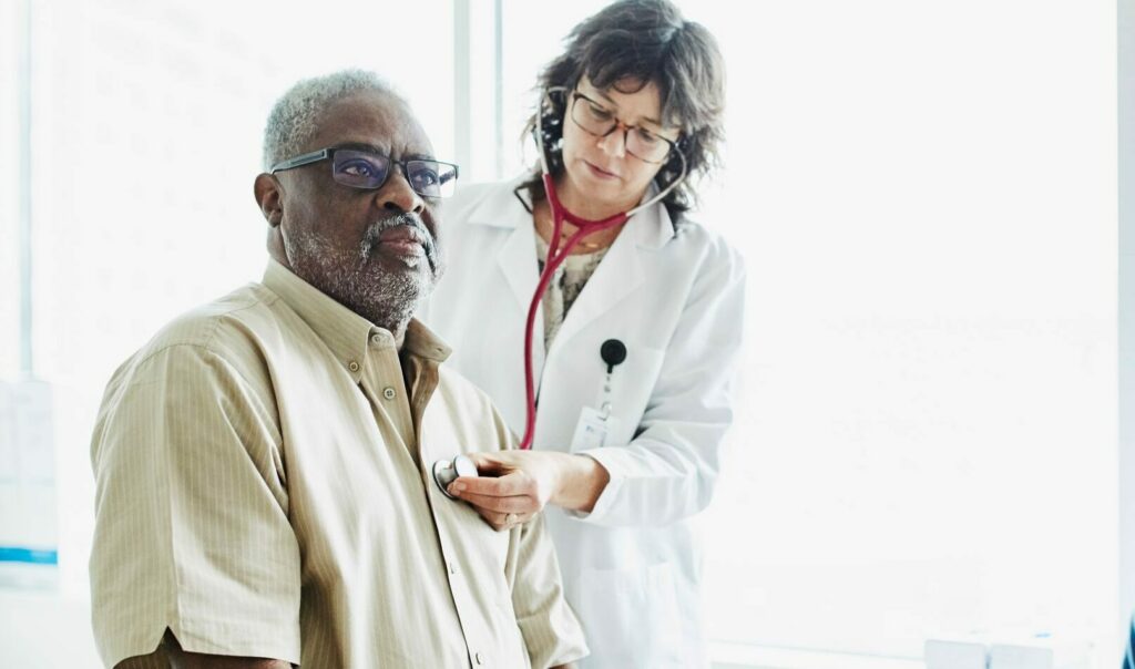 Female doctor listening to an older man's heart while a younger woman watches.