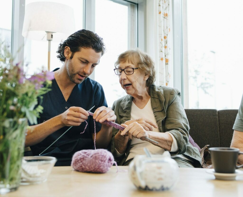 Retired senior woman teaching knitting to male nurse while sitting on sofa at elderly care home